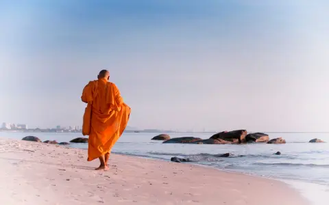 A modern day monk in an orange robe walks along a serene beach at sunrise, with calm ocean waves and distant city skyline.