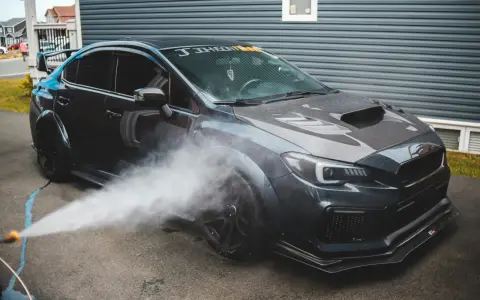 Nettoyage d'une voiture de sport sombre. Un jet d'eau est dirigé vers la roue avant, peut-être pour enlever mousse sur goudron vinaigre blanc.