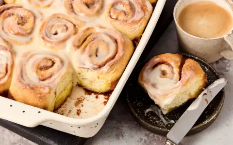 Saftige, vegane Zimtschnecken mit cremiger Glasur in einer Auflaufform, daneben eine Tasse Kaffee und eine einzelne Schnecke auf einem Teller.