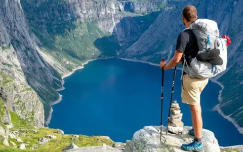 Un escursionista con zaino e bastoncini ammira un lago blu intenso circondato da montagne imponenti. Un'avventura di trekking montagna indimenticabile.