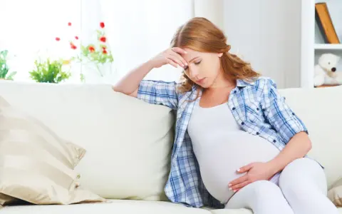 Zmęczona kobieta w 26 tygodniu ciąży siedzi na kanapie, trzymając się za głowę.