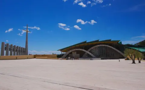 The modern church of San Giovanni Rotondo Padre Pio stands under a bright blue sky, with a tall cross and bell tower in the distance.