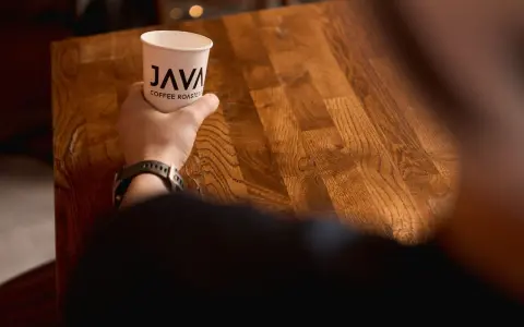 Dłoń trzyma kubek z logo "JAVA COFFEE ROASTERS", gotowa do degustacji świeżego batch brew na drewnianym stole.