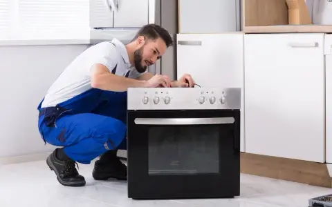 Un technicien en salopette bleue répare une prise four. Il est accroupi devant l'appareil, concentré sur les fils.