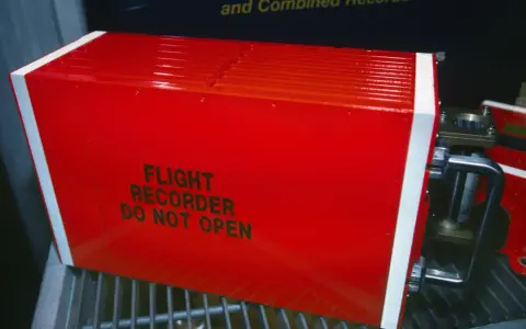 Czerwony, jaskrawy rejestrator lotu, znany jako czarna skrzynka inaczej, z napisem "FLIGHT RECORDER DO NOT OPEN".