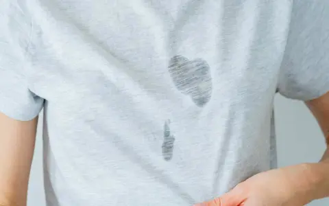 Gros plan sur une tache d'huile sur un vêtement gris clair. La tache est sombre et semble avoir pénétré le tissu.