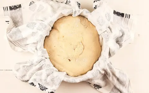 Masa de pan de patata cruda, cubierta con un paño blanco con estampado floral gris, lista para levar.