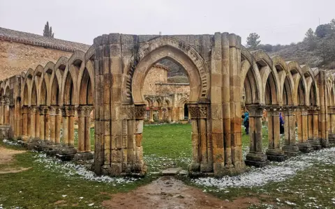 Ruinas de un claustro con arcos de piedra, evocando el nacimiento del Duero. Nieve ligera cubre el césped.