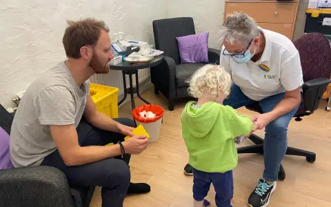 En pappa och ett barn får vaccin av en sjuksköterska. Barnet verkar lite oroligt, men sjuksköterskan är lugn och trygg. Det är en fin stund av vaccin italien.