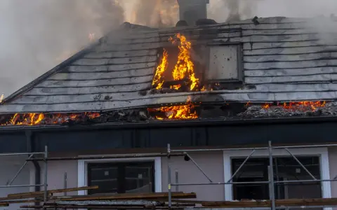 Un **incendie habitation** ravage le toit d'une maison, avec des flammes vives et une épaisse fumée s'échappant des combles.