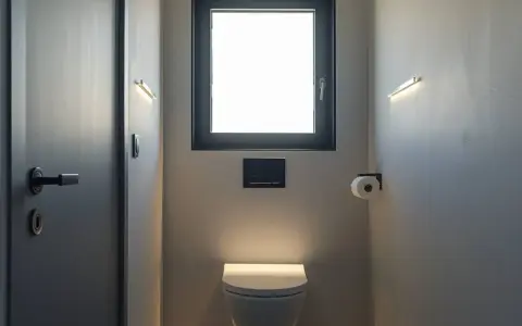 Une toilette moderne avec une fenêtre au-dessus, éclairée par des appliques murales discrètes. Un rouleau de papier toilette est à portée de main.