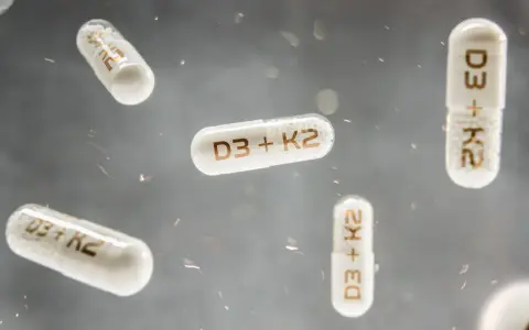 Flera kapslar med texten "D3 + K2" svävar i luften. Dessa kapslar kan vara ett komplement till din k vitamin mat.