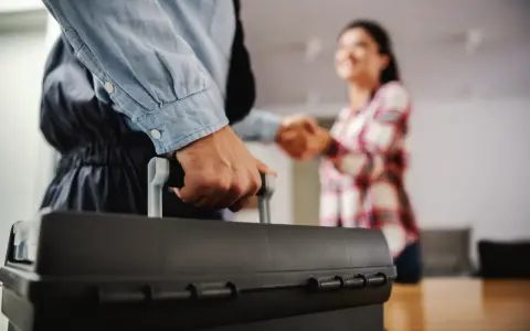 Un plombier arrive avec sa mallette pour régler une fuite d'eau avant compteur. Une cliente le remercie.