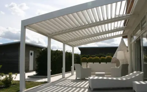 Un moderno porche de obra con techo de lamas ajustables, mobiliario de exterior blanco y un camino de madera que conduce a una cabaña.