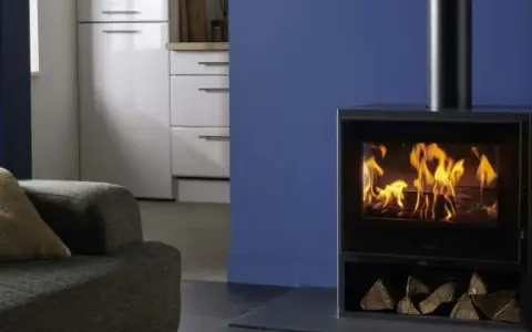Estufa de leña moderna con fuego encendido y leña apilada, junto a un sofá y muebles de cocina.