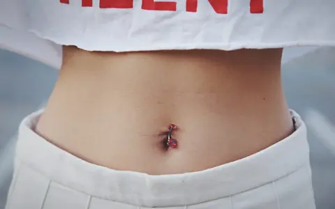 Brzuszek z kolczykiem w pępku. Czy dziurka w pępku zarasta? Delikatna skóra, białe spodenki i crop top z czerwonym napisem.