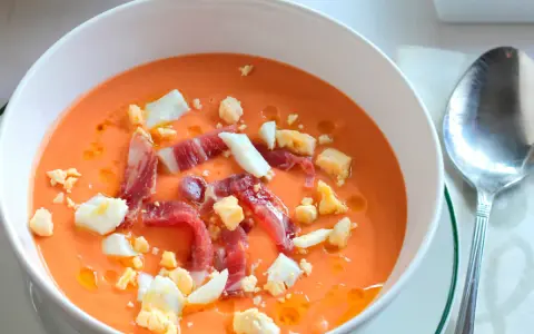 Un cuenco blanco con salmorejo cordobés, coronado con huevo duro picado y taquitos de jamón serrano.