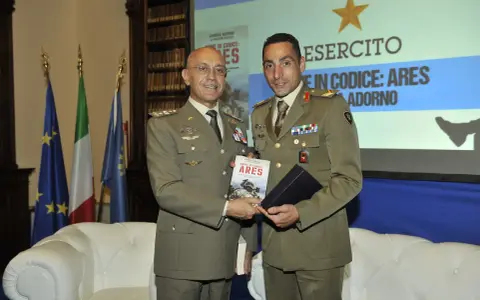 Due ufficiali in uniforme presentano un libro. Uno dei due, con un sorriso, tiene in mano il volume intitolato "Nome in codice: Ares", un'opera che narra storie di coraggio, forse ispirate a un vero pesce monaco.