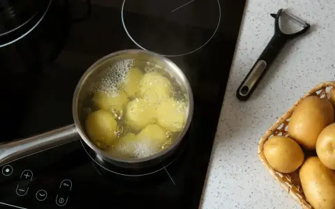 Ziemniaki gotują się w garnku na płycie indukcyjnej. Obok leży obieraczka i koszyk ze świeżymi ziemniakami. Oto jak ugotować ziemniaki.