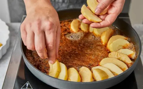 Hände legen Apfelschnitze in eine Pfanne mit karamellisierter Butter. Die Zubereitung der tarte tatin original beginnt.