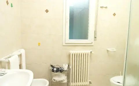Bagno 79 Rimini: lavabo, bidet, WC, scaldasalviette e finestra. Asciugacapelli e rotolo di carta igienica su carrello.