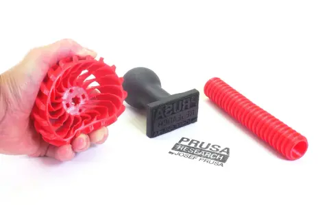 Czerwony, elastyczny filament w kształcie wirnika, stempel z logo Prusa Research i czerwona rurka.