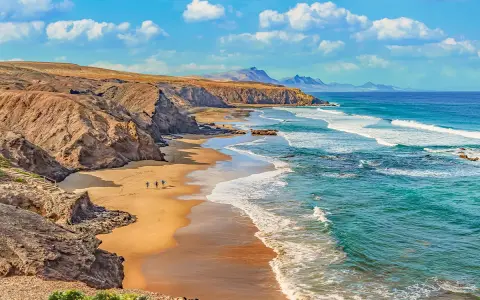 Plaża z klifami i turkusowym oceanem, kilka osób z deskami surfingowymi idzie po piasku.