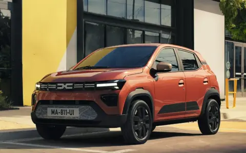 Pomarańczowy Dacia Spring stoi przed budynkiem z żółtą ścianą.