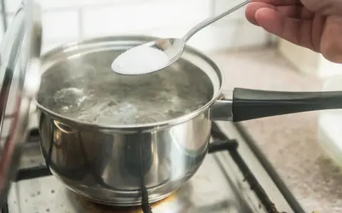 En sked med salt tillsätts i kokande vatten. En del av locket är upplyft. En burk med salt syns i bakgrunden, redo för egen resorb.