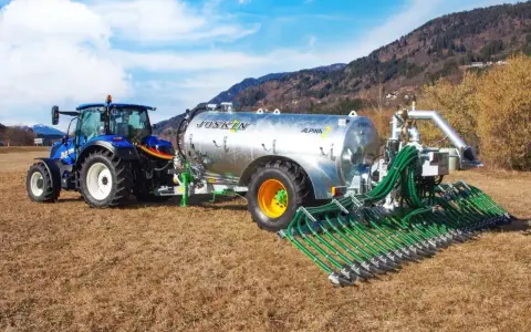 Niebieski traktor ciągnie przyczepę z cysterną do przewozu gnojowicy, wyposażoną w rozdzielacz z wieloma wężami do precyzyjnego nawożenia pól.