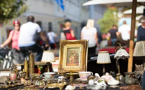 Antikviteter och kuriosa på ett bord, kanske på en loppmarknad i Stockholm. En blomstermålning i guldram, små lampor och porslin.
