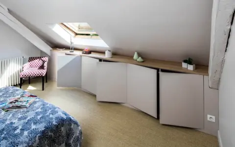 Chambre mansardée d'un coté avec lit, fauteuil à pois et rangements bas sur mesure. Un velux éclaire la pièce.