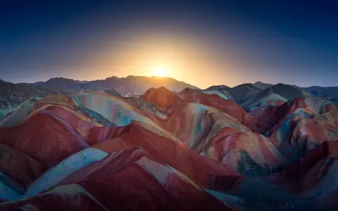 Bunte Schichten aus Rot, Gelb und Blau formen die **Regenbogenberge Chinas** unter einem tiefblauen Himmel mit aufgehender Sonne.