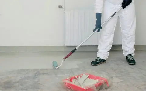 Person i skyddsdräkt målar betonggolv med en roller. Färgen är ljus och en färgtråg syns på golvet.