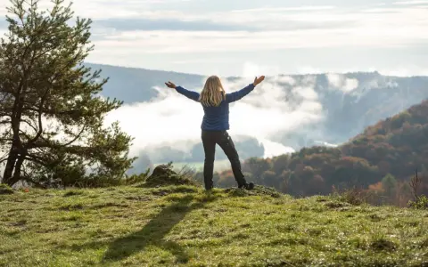 Frau mit ausgebreiteten Armen auf einem Hügel, umgeben von Nebel und Herbstfarben. Das Gefühl von Freiheit und die Bedeutung des Pilgerns werden hier spürbar.