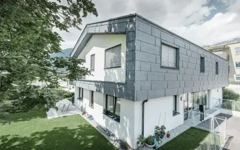 Esterno di una casa grigia moderna con tetto spiovente e facciata in ardesia. Un giardino verde e alberi ombreggiano la proprietà.