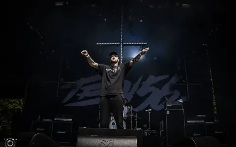 Un rappeur tatoué, casquette et lunettes de soleil, bras levés sur scène, devant un fond noir avec le logo "TEN 56".