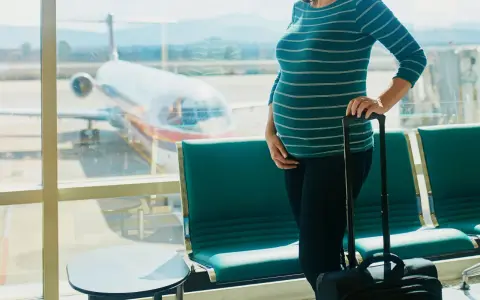 Ciężarna kobieta z walizką czeka na lotnisku. Widok na samolot przez okno. Latanie samolotem w ciąży to wyzwanie, ale podróż trwa.