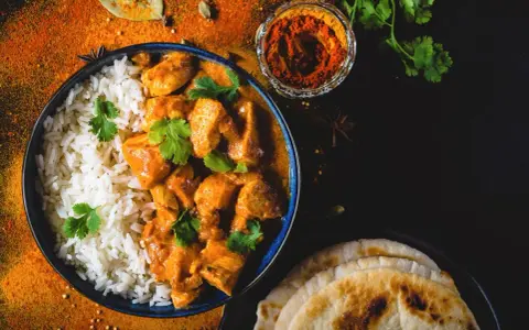 Pyszny Butter Chicken przepis: kremowy kurczak w sosie pomidorowym z ryżem i plackami naan, ozdobiony świeżą kolendrą i przyprawami.