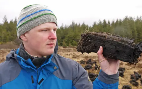 Mann mit Mütze hält Torfstück, das an rauchige Whiskys erinnert. Wald im Hintergrund.