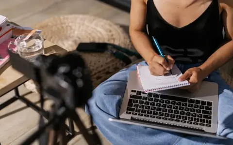 Kobieta pisze notatki w zeszycie, przygotowując materiał na artykuł. Obok leży laptop i szklanka wody.