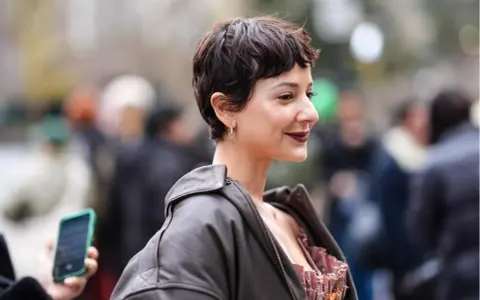 Frau mit einem lässigen Pixie-Cut, dunklem Lippenstift und Lederjacke lächelt in die Kamera.