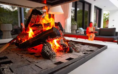 En kamin som sprakar med livliga lågor. En kvinna sitter i bakgrunden och läser, njuter av värmen. En perfekt kväll att elda i kamin varje dag.