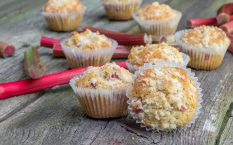 Pyszne muffinki z rabarbarem, posypane kruszonką, leżą na drewnianym blacie obok świeżych łodyg rabarbaru.