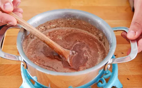 Hände rühren eine schokoladige Masse in einem Topf. So einfach lässt sich die Glasur selber machen für Kuchen oder Gebäck.