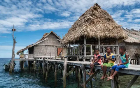 Trzech chłopców siedzi na drewnianym pomoście przy tradycyjnych chatach na wodzie w Raja Ampat.