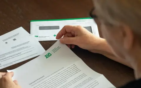 Dłoń starszej osoby przegląda dokumenty z ZUS. W tle koperta z logo ZUS.