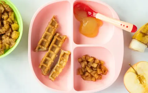 Baby-freundliche Apfelwaffeln mit Apfelmus und Zimt-Apfelstückchen auf einem rosa Teller. Ein halber Apfel und eine Banane liegen daneben.
