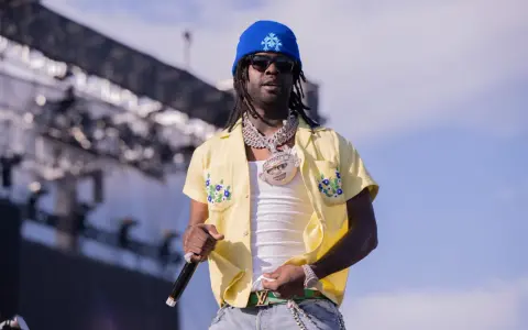 Chief Keef, w niebieskiej czapce i żółtej koszuli z kwiatami, na scenie. Jego wiek nie jest widoczny, ale styl mówi wszystko.