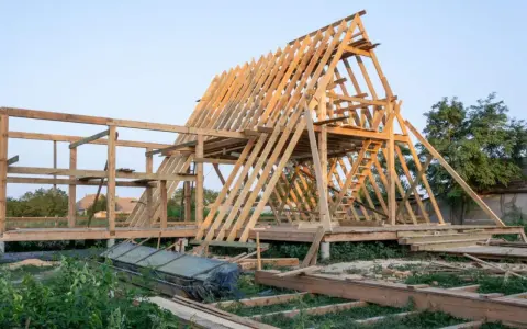 Drewniana konstrukcja domu w budowie. Widoczny szkielet dachu i ścian, schody. Fundament punktowy pod domek drewniany jest już gotowy.